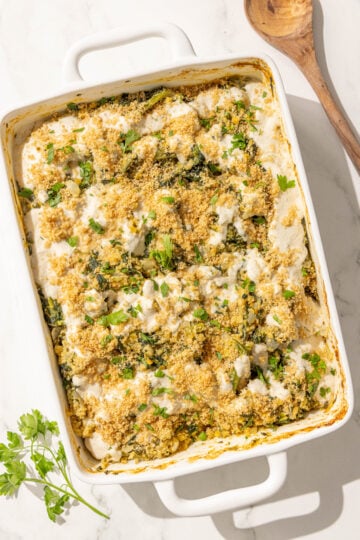 A 9x13 spinach cheesy quinoa casserole topped with parsley and breadcrumbs and a wooden spoon on the counter next to the dish.