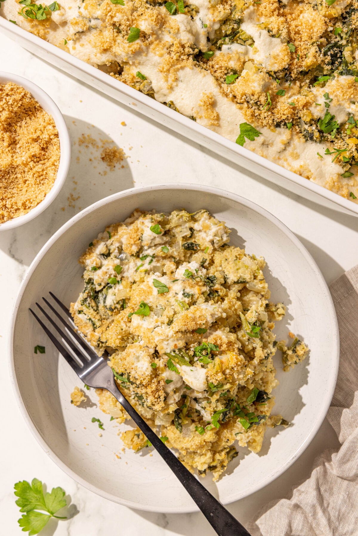 A bowl of quinoa spinach casserole with a fork in it, a casserole dish with remaining casserole, and a small bowl of breadcrumbs.