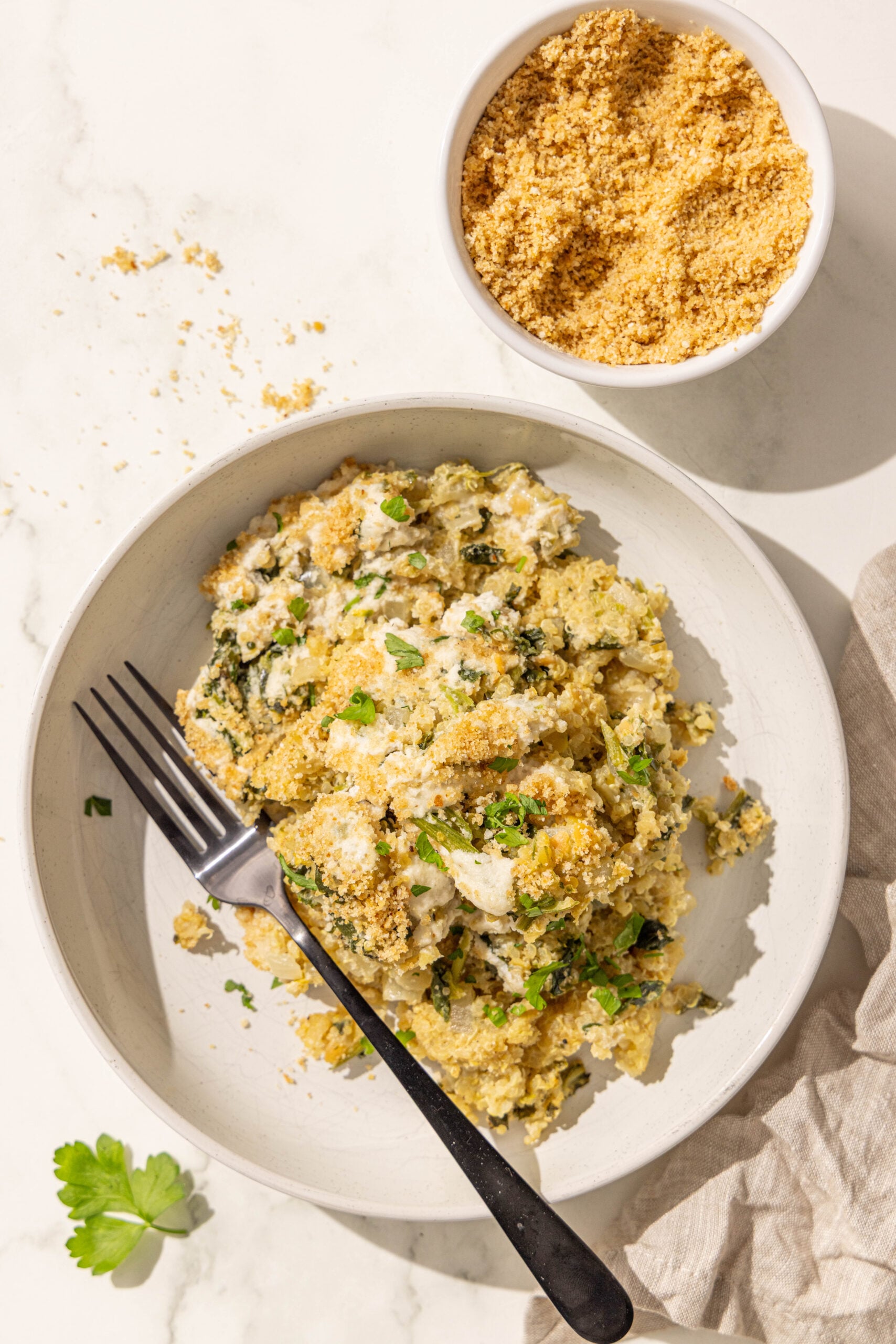 A bowl of spinach quinoa casserole with a fork in it and a small bowl of breadcrumbs.