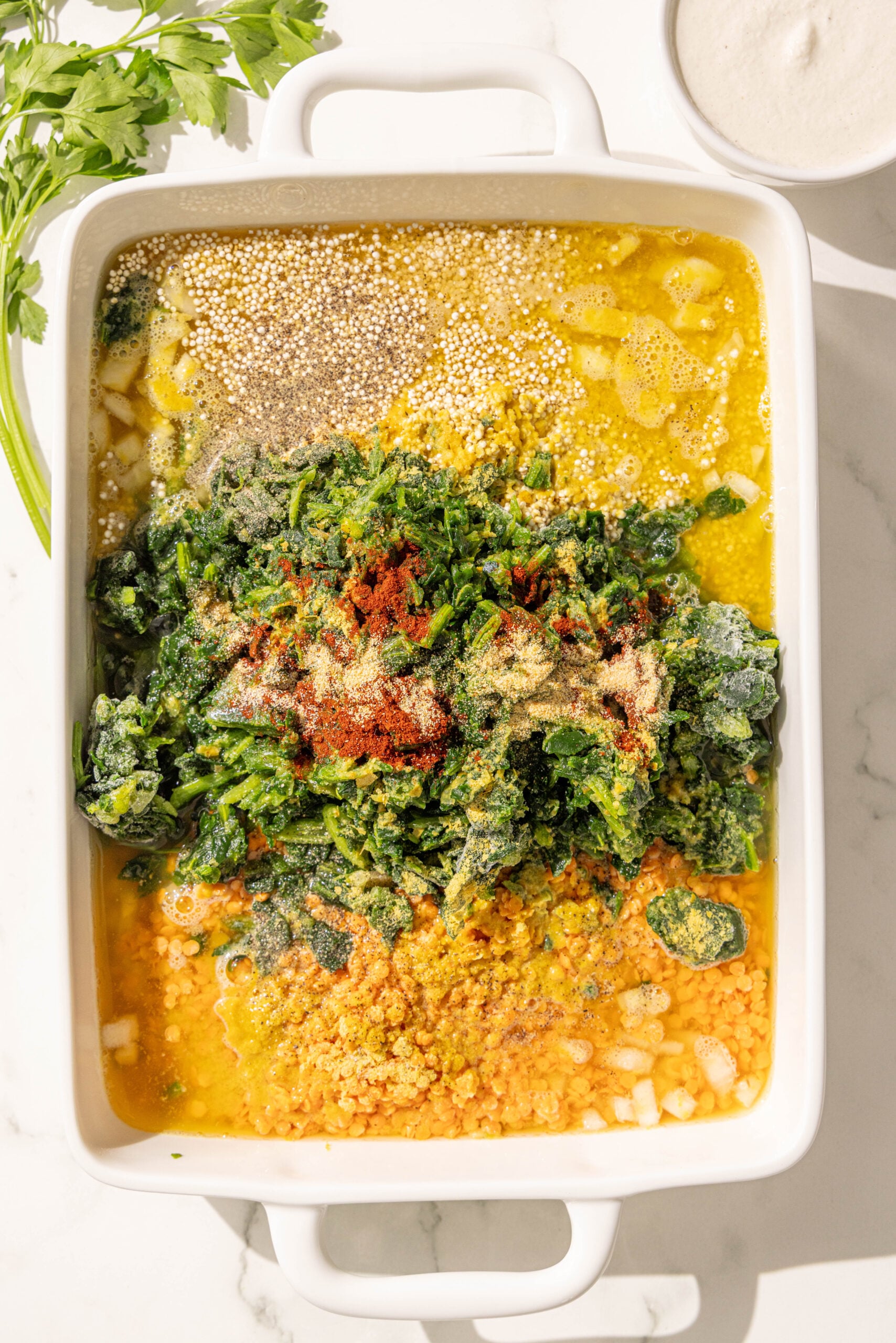 A large 9x13 white casserole dish with quinoa, red lentils, onion, spinach, vegetable broth, and spices in it.