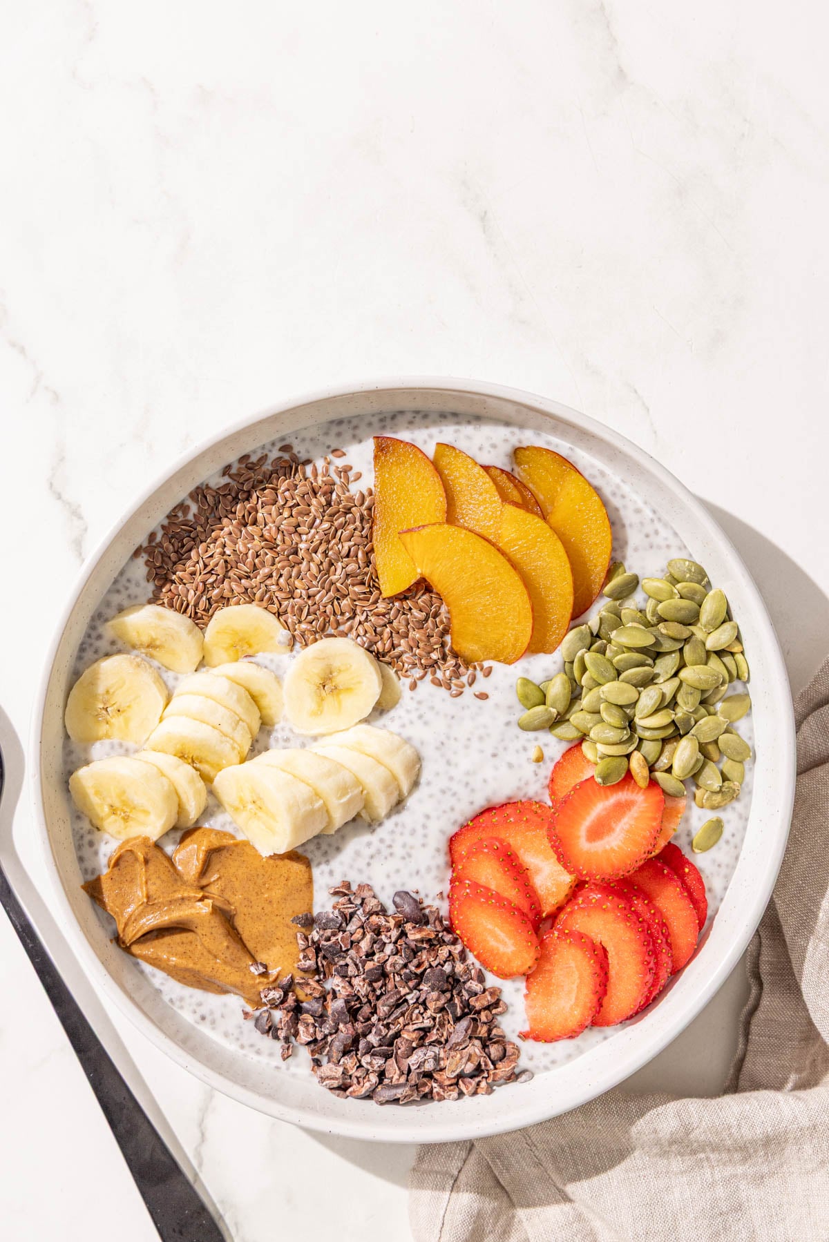 A bowl of chia pudding topped with flax seed, sliced plum, strawberries, banana, pumpkin seeds, almond butter, and cacao nibs plus a black metal spoon.