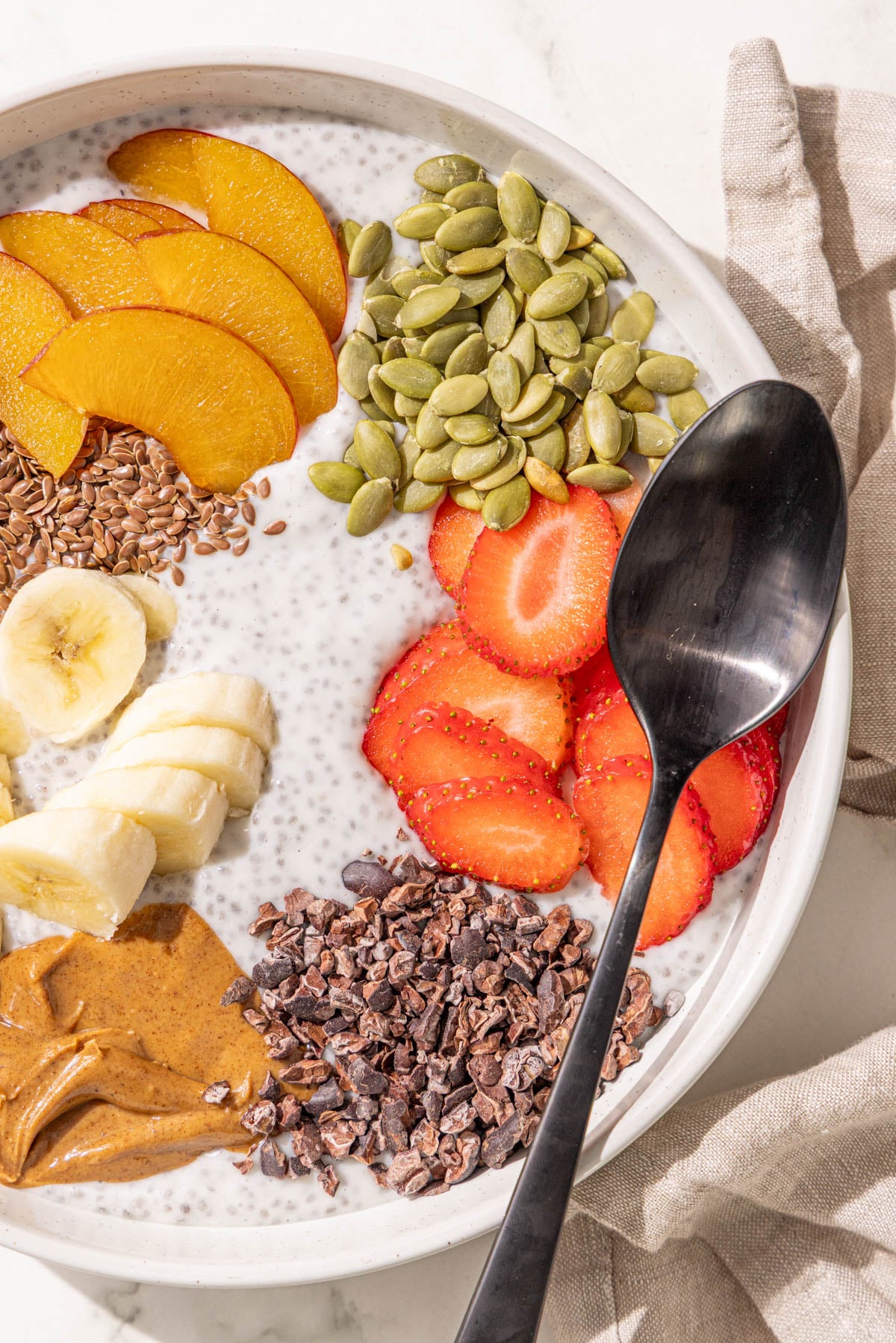 A bowl of chia pudding topped with flax seed, sliced plum, strawberries, banana, pumpkin seeds, almond butter, and cacao nibs with a black metal spoon in it.