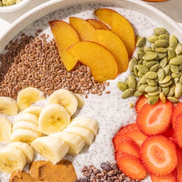 A bowl of chia pudding topped with flax seed, sliced plum, strawberries, banana, pumpkin seeds, almond butter, and cacao nibs.