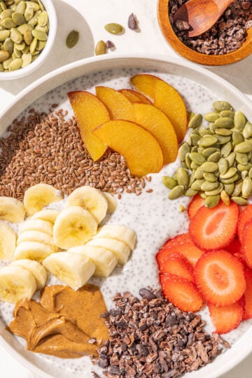 A bowl of chia pudding topped with flax seed, sliced plum, strawberries, banana, pumpkin seeds, almond butter, and cacao nibs.