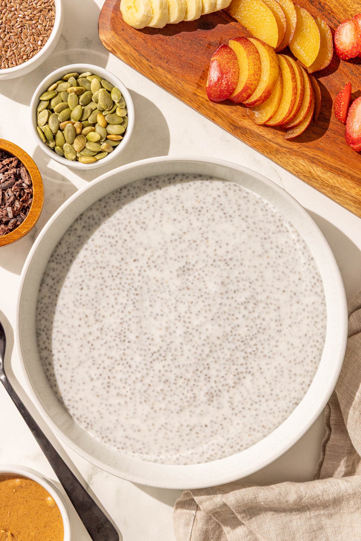 A bowl of chia pudding and smaller bowls of pumpkin seeds, cacao nibs, flax seeds, almond butter, and a cutting board with sliced plum and banana on it.
