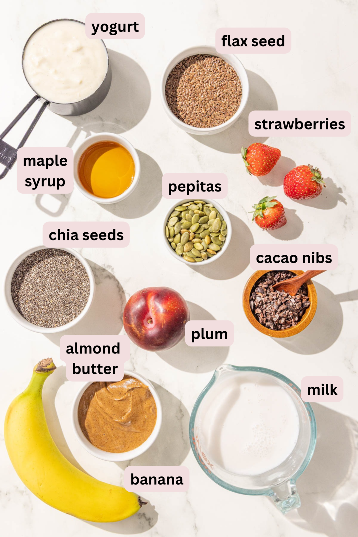 Ingredients laid out in bowls and labeled including yogurt, flax seed, maple syrup, pepitas, strawberries, chia seeds, cacao nibs, plum, almond butter, banana, and milk.
