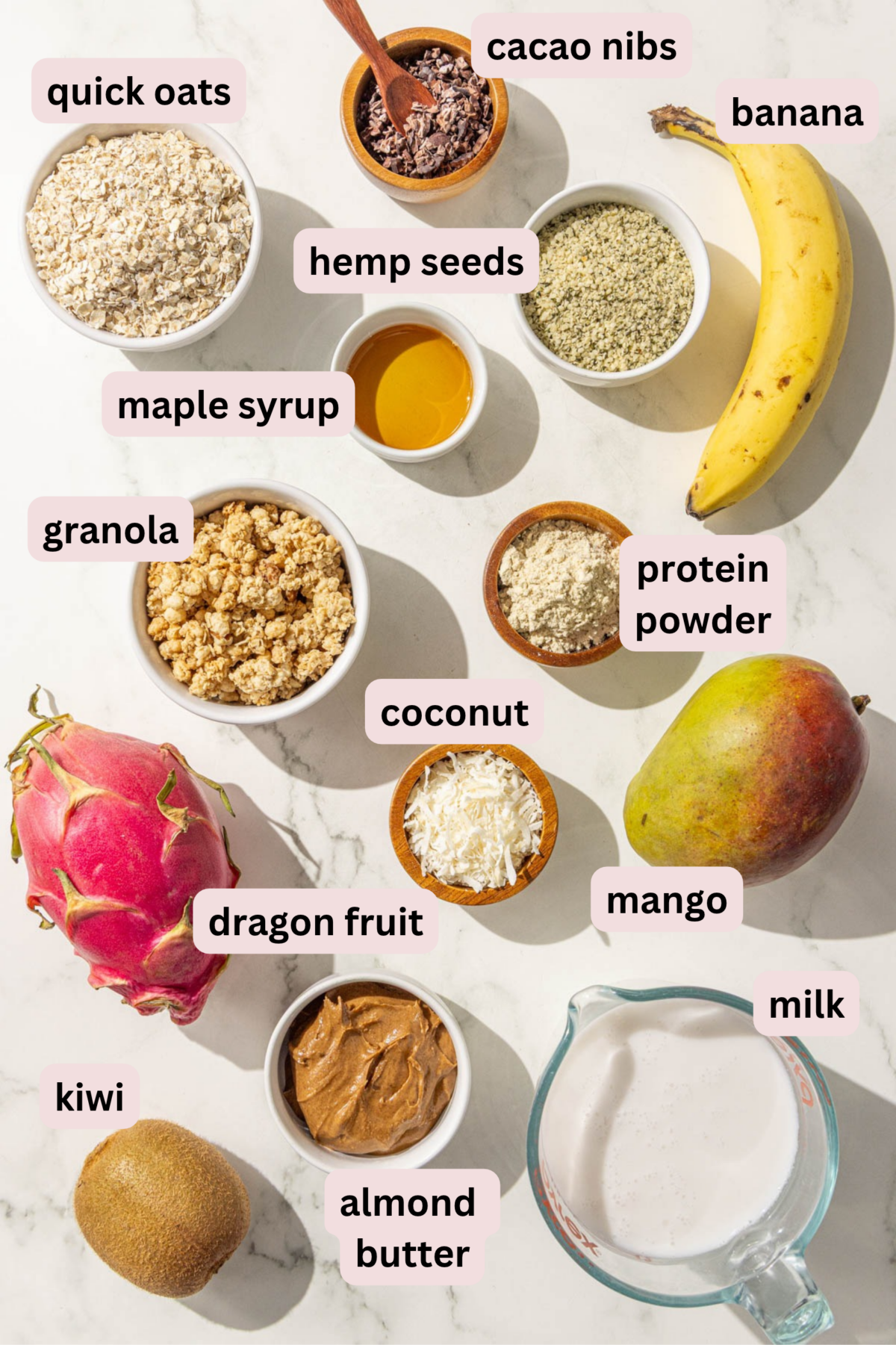 Ingredients laid out on a marble countertop, in bowls, and labeled including: quick oats, cacao nibs, a whole banana, hemp seeds, maple syrup, granola, protein powder, a whole mango, shredded coconut, a whole dragon fruit, milk, almond butter, and a whole kiwi.