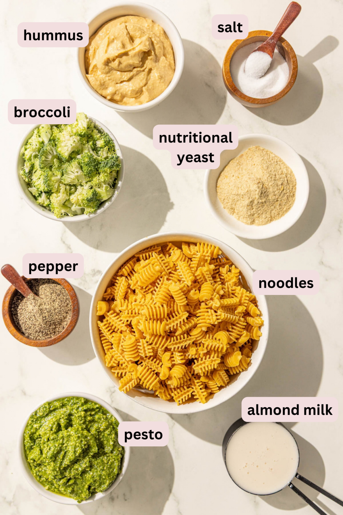 Ingredients laid out on a marble countertop in bowls and labeled including hummus, salt, broccoli, nutritional yeast, noodles, pepper, almond milk, and pesto.