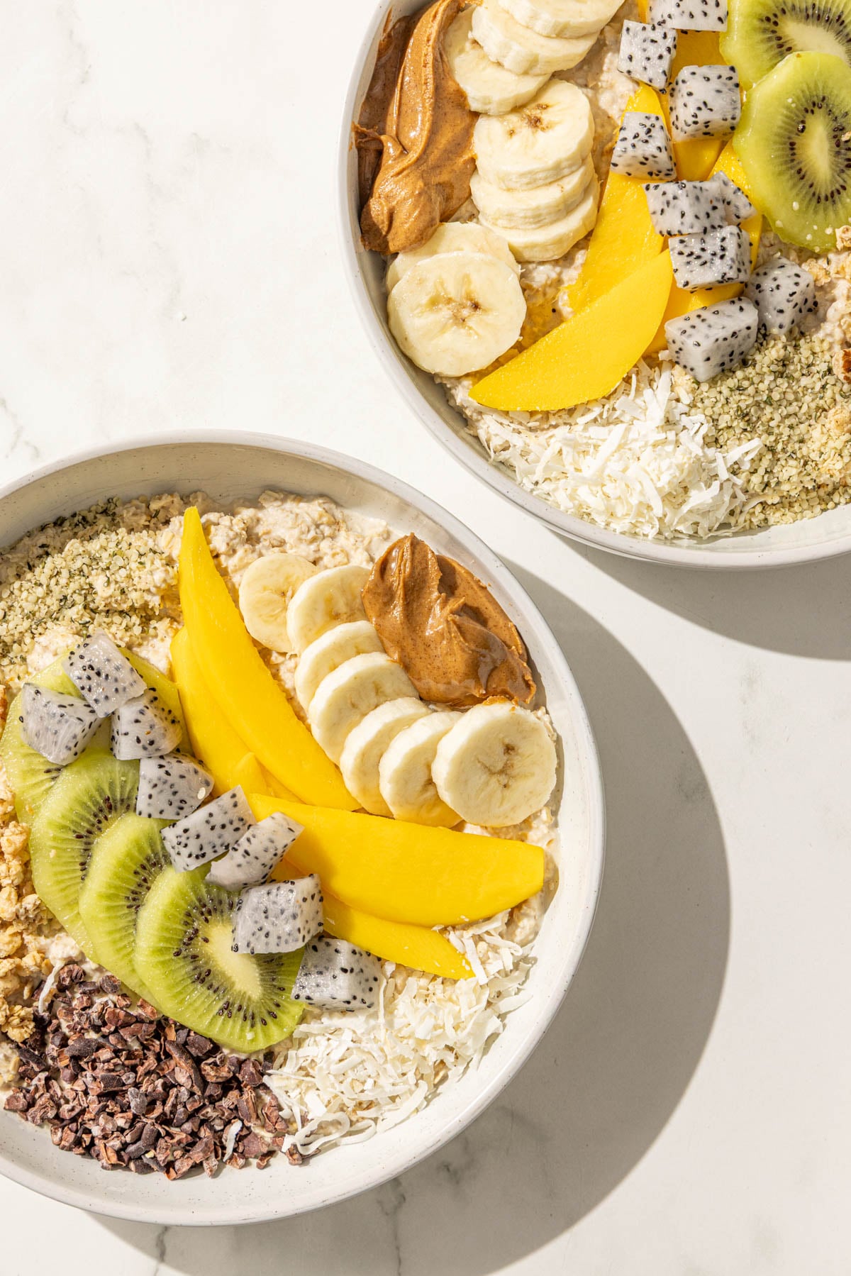 Two bowls of oatmeal topped with kiwi, banana, mango, dragon fruit, cacao nibs, coconut, granola, hemp seeds and almond butter.