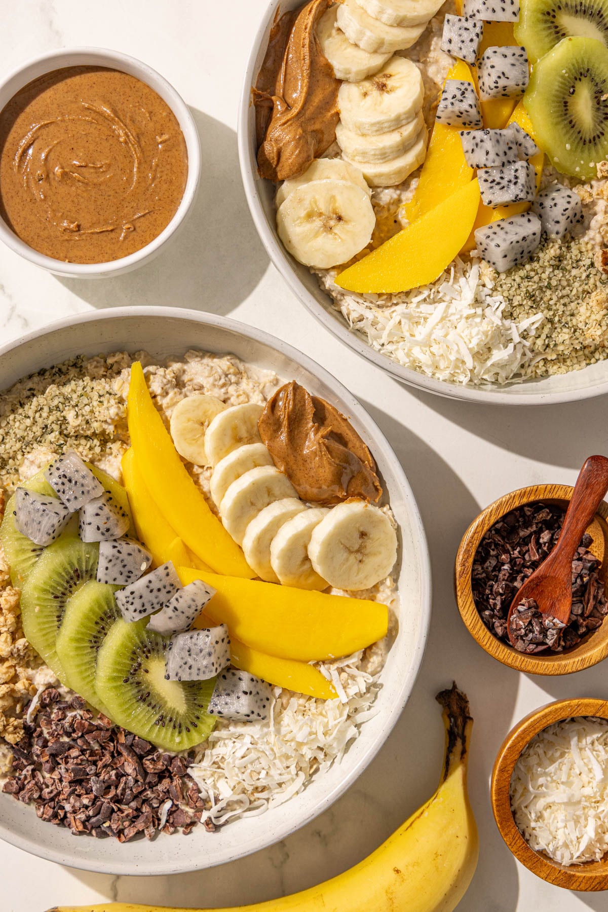 Two bowls of oatmeal topped with kiwi, banana, mango, dragon fruit, cacao nibs, coconut, granola, hempseeds and almond butter. There is also a bowl of almond butter, a bowl of cacao nibs, a bowl of shredded coconut, and a whole banana.