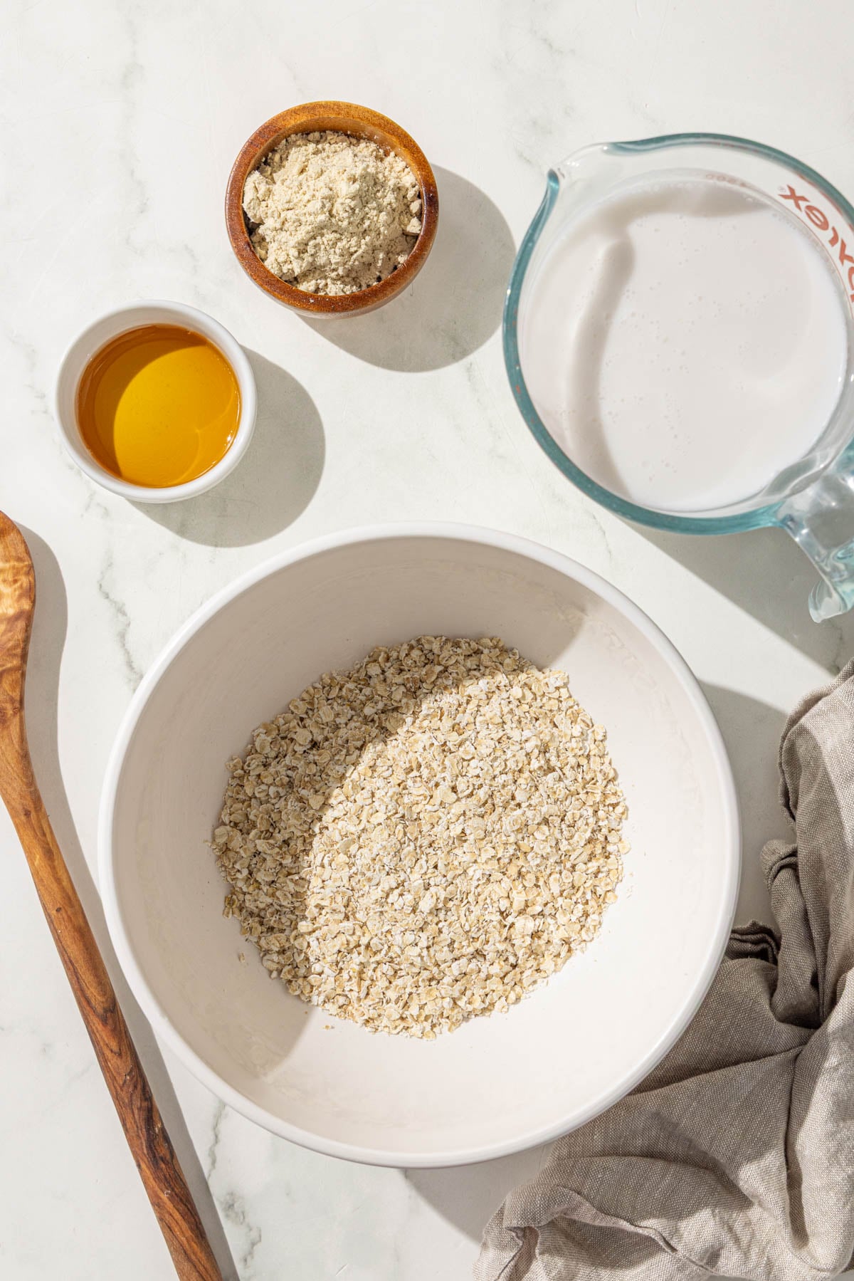 A bowl of quick oats, a small bowl of maple syrup, a small bowl of protein powder, a glass jar with milk in it and a wooden spoon.