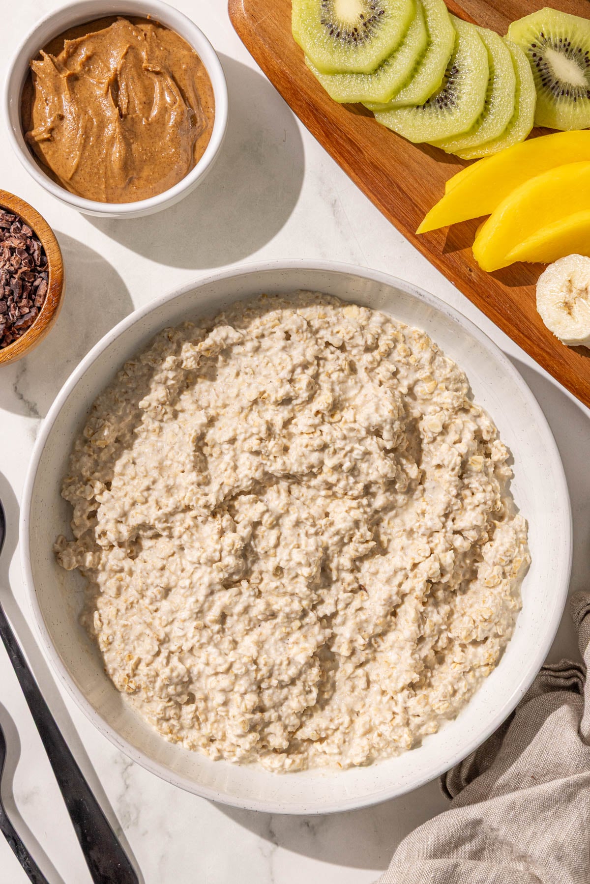 A bowl of oatmeal surrounded by a small bowl of almond butter, cacao nibs, two black metal spoons, and a wooden cutting board with sliced kiwi, banana, and mango on it.