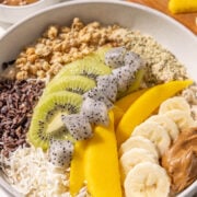 A bowl of oatmeal topped with kiwi, banana, mango, dragon fruit, cacao nibs, coconut, granola, hempseeds and almond butter. There is also a bowl of almond butter and a wooden cutting board with kiwi, mango, and dragonfruit on it.