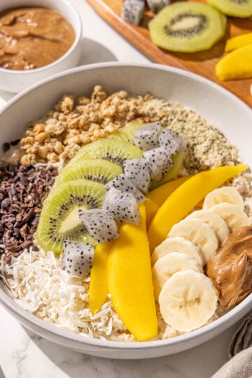 A bowl of oatmeal topped with kiwi, banana, mango, dragon fruit, cacao nibs, coconut, granola, hempseeds and almond butter. There is also a bowl of almond butter and a wooden cutting board with kiwi, mango, and dragonfruit on it.