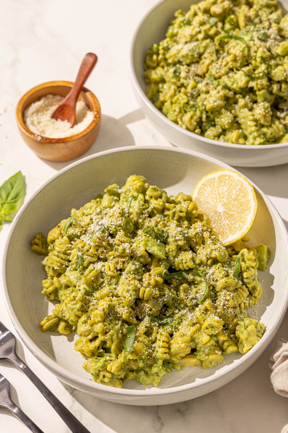 Two bowls of hummus pesto broccoli pasta, a small wooden bowl of parmesan, and two black metal spoons.