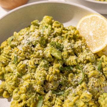 A bowl of hummus pesto broccoli pasta with a lemon wedge in the bowl and a bowl of grated parmesan in the background.