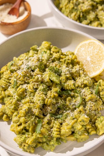 A bowl of hummus pesto broccoli pasta with a lemon wedge in the bowl and a bowl of grated parmesan in the background.