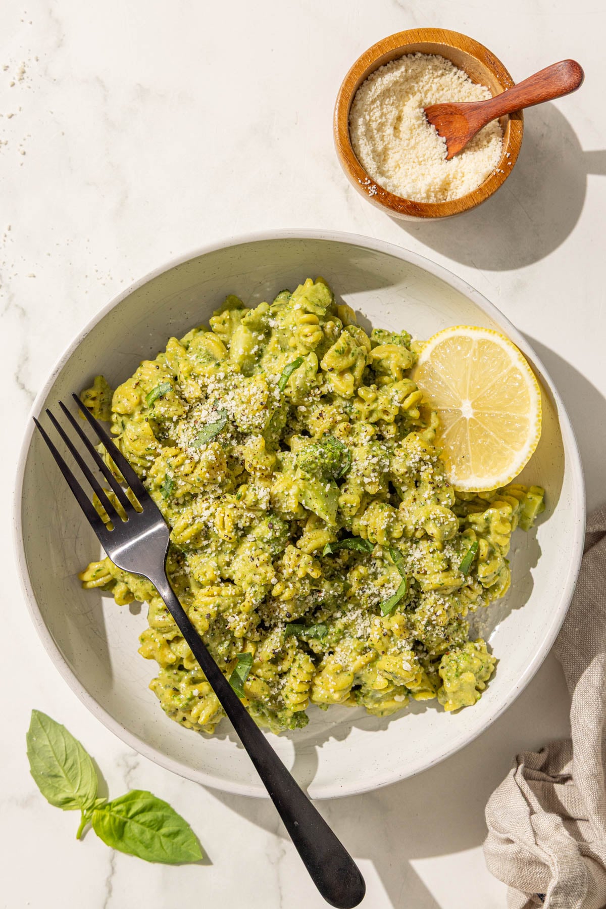 A bowl of pesto hummus broccoli pasta topped with parmesan and a lemon wedge with a black fork in it and a small bowl of parmesan beside the bowl.