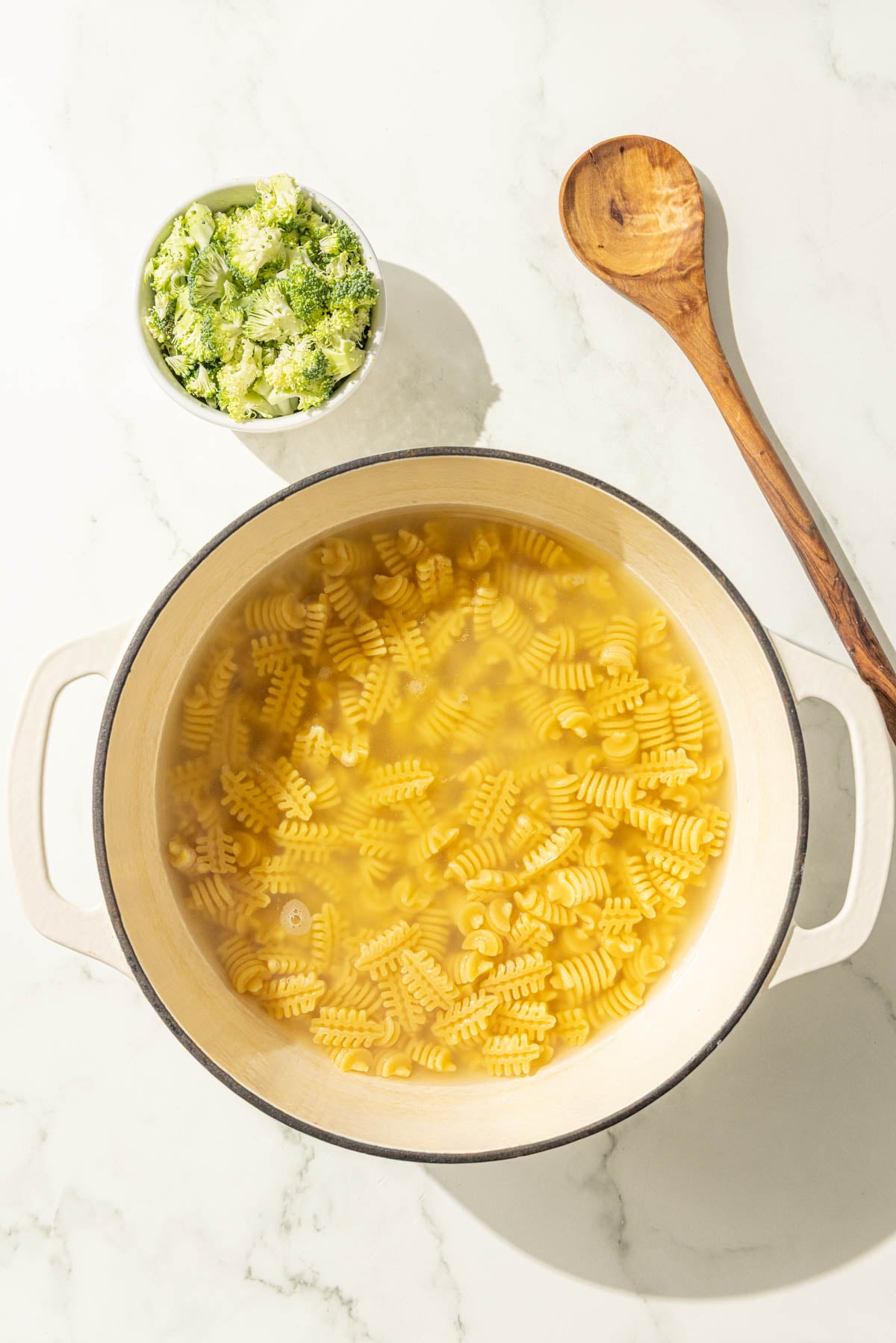 A large pot of noodles in water, a small bowl of broccoli, and a wooden spoon.