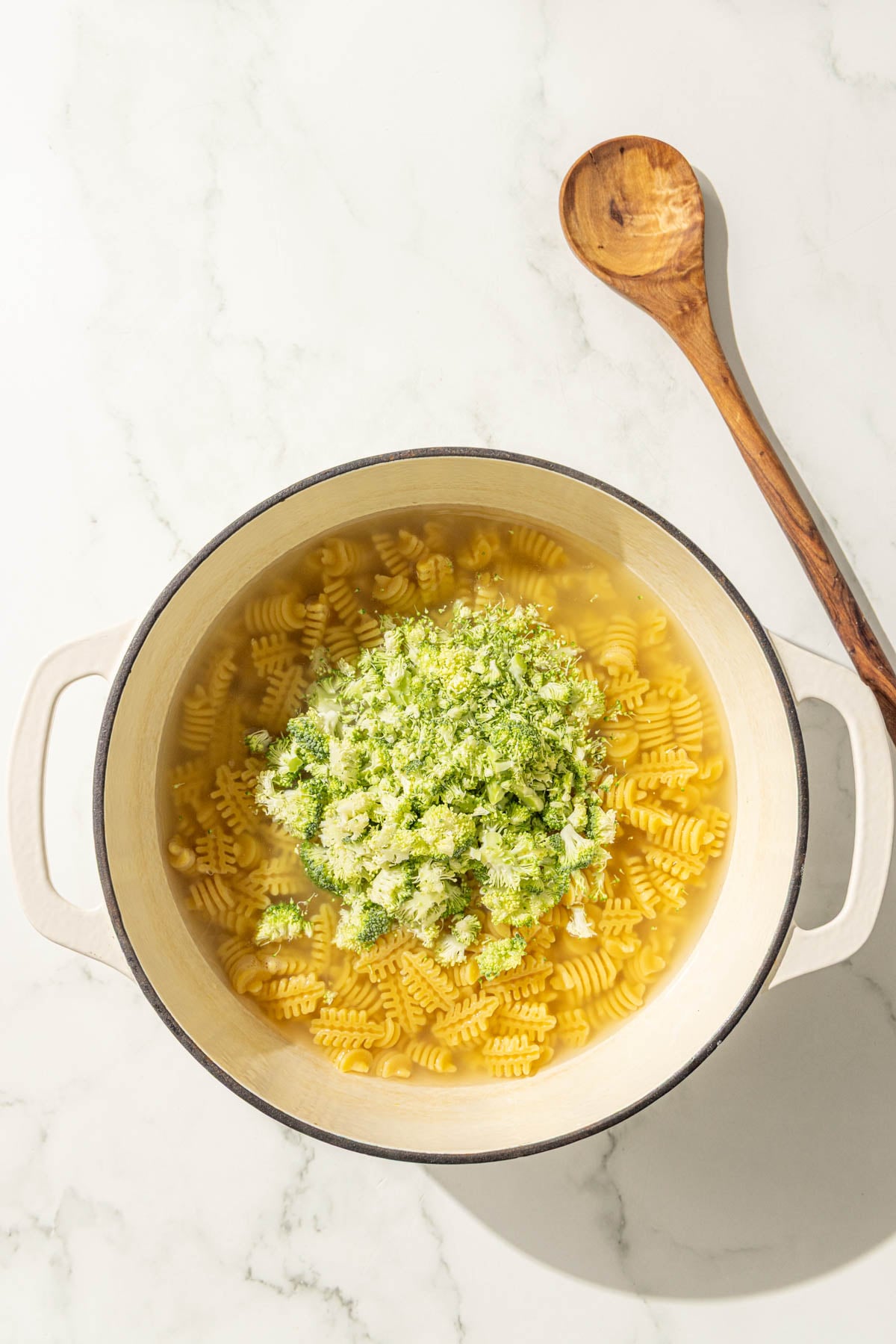 A large pot of noodles and broccoli in water and a wooden spoon.