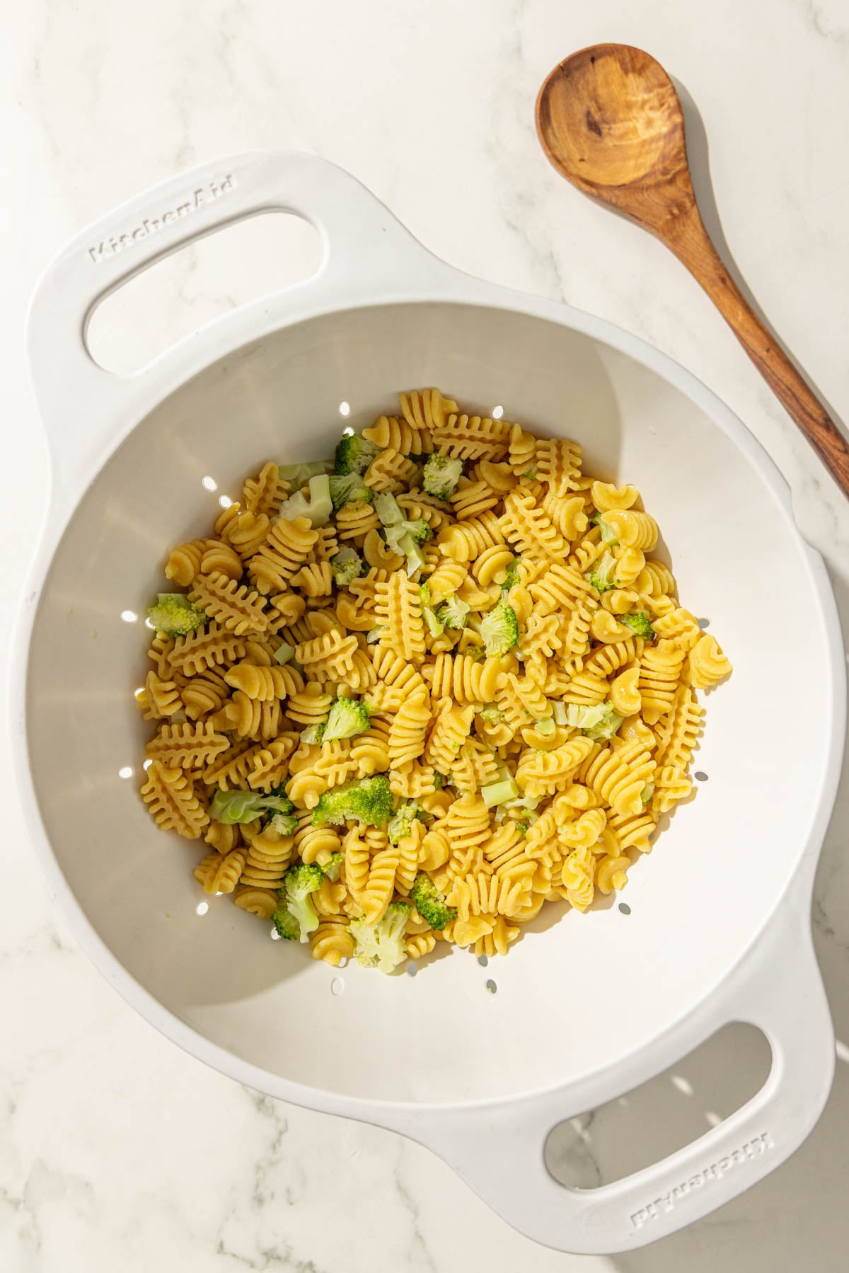 A strainer filled with noodles and broccoli and a wooden spoon.