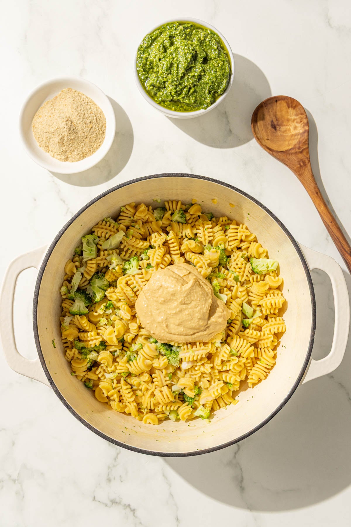 A large pot of cooked noodles and broccoli with a dollop of hummus in it along with a bowl of pesto, a bowl of nutritional yeast, and a wooden spoon.